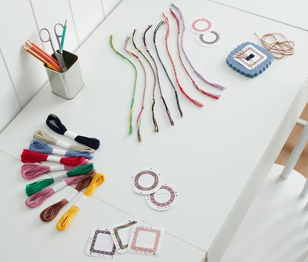 Sur une table blanche sont posés un kit de bricolage « bracelets brésiliens », des fils colorés et une lampe bleue.