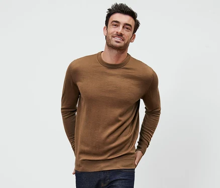 Un homme pose sur un fond blanc et porte un pull en mérinos cognac.