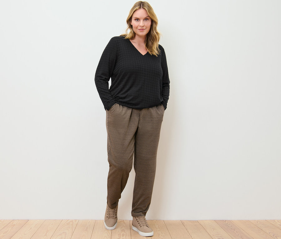 Une femme se tient debout, les mains dans les poches d'un pantalon de jogging avec jacquard pied-de-poule et d'un t-shirt manches longues structuré noir.