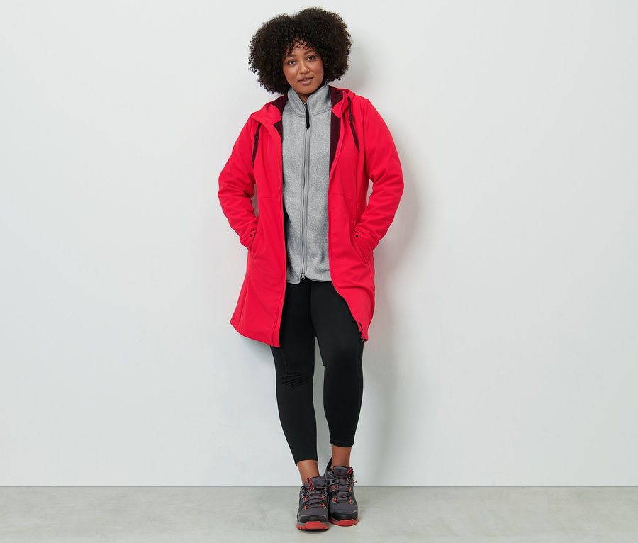 Une femme porte un manteau softshell rouge, une veste en maille polaire gris clair et un legging thermique.