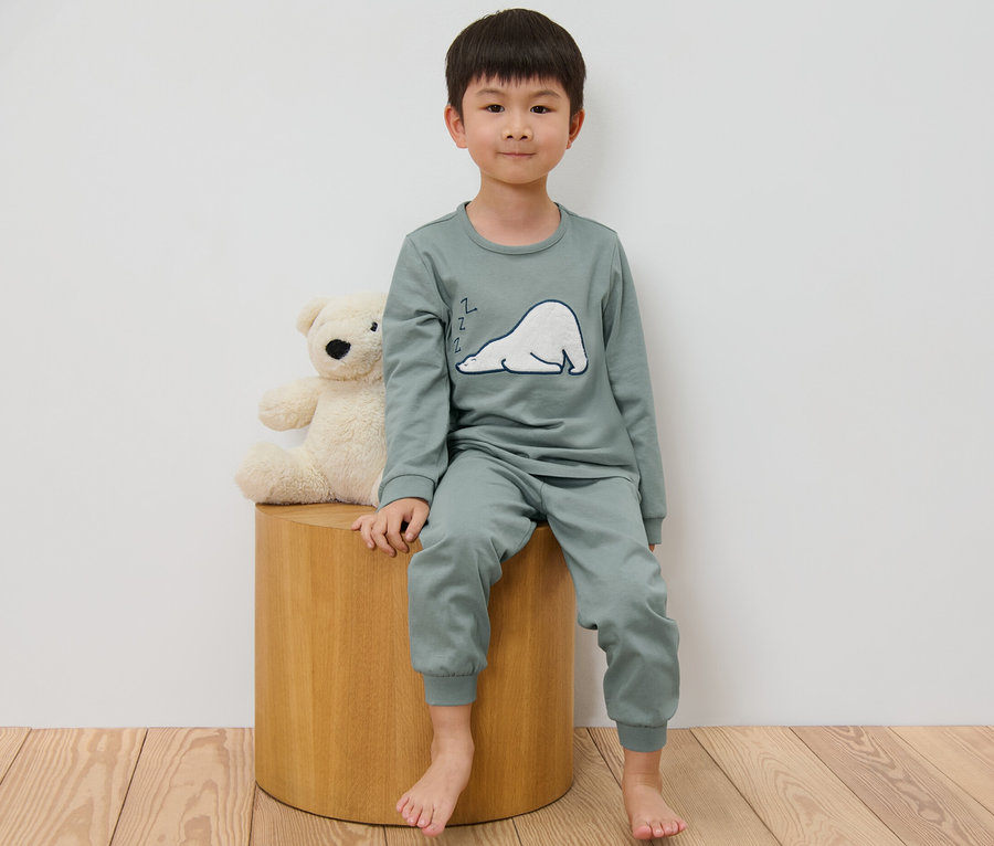 Un garçon est assis sur un tabouret à côté d'un ours en peluche et porte un pyjama pour enfant.