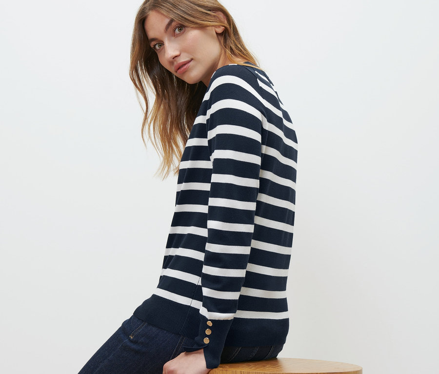 Une femme est assise sur un tabouret en bois et porte un pull en tricot fin à rayures bleu marine et un jean.