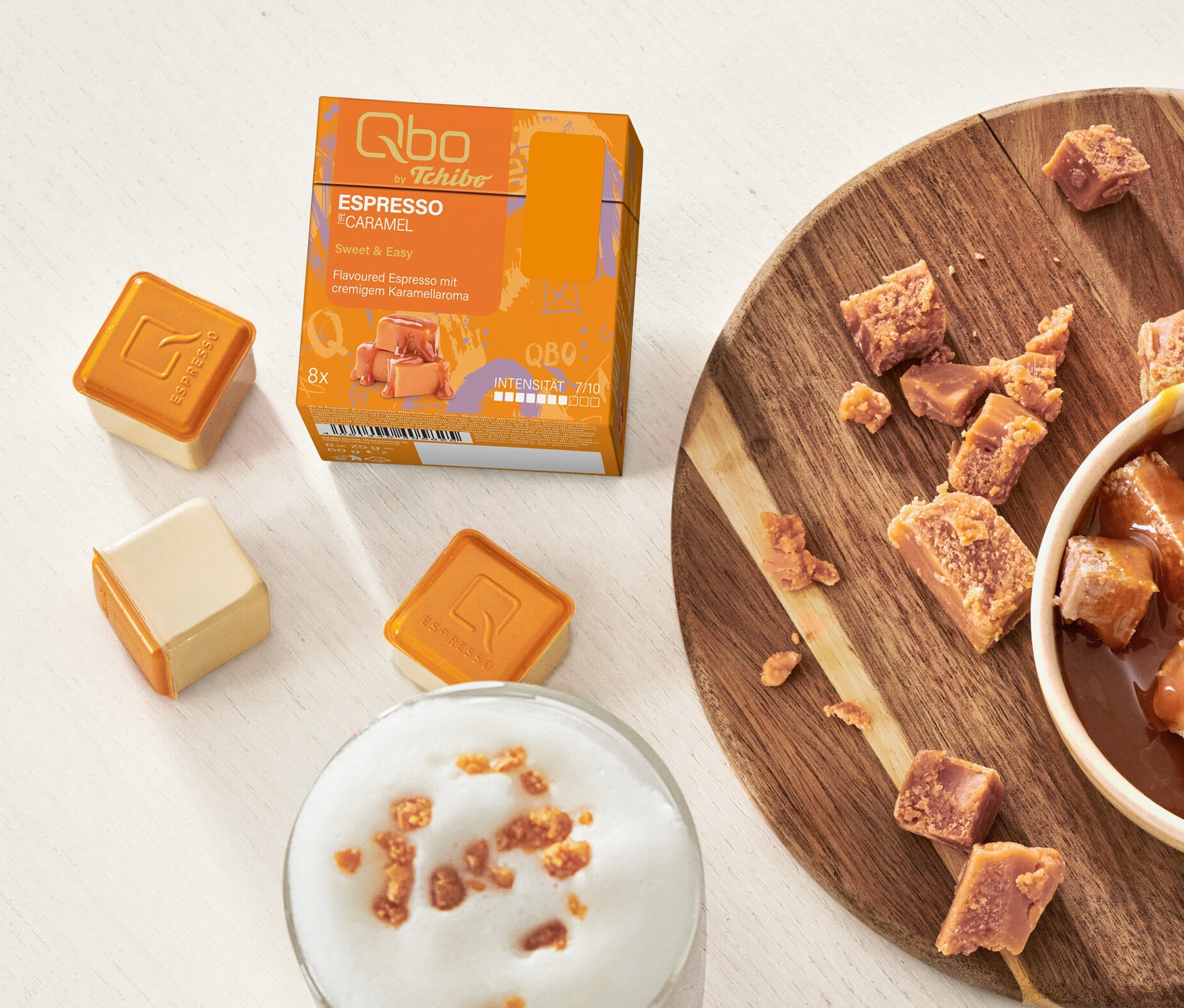 Sur une table, il y a des capsules Qbo Flavoured Espresso Caramel, un bol de cubes de caramel et un verre de latte au caramel.