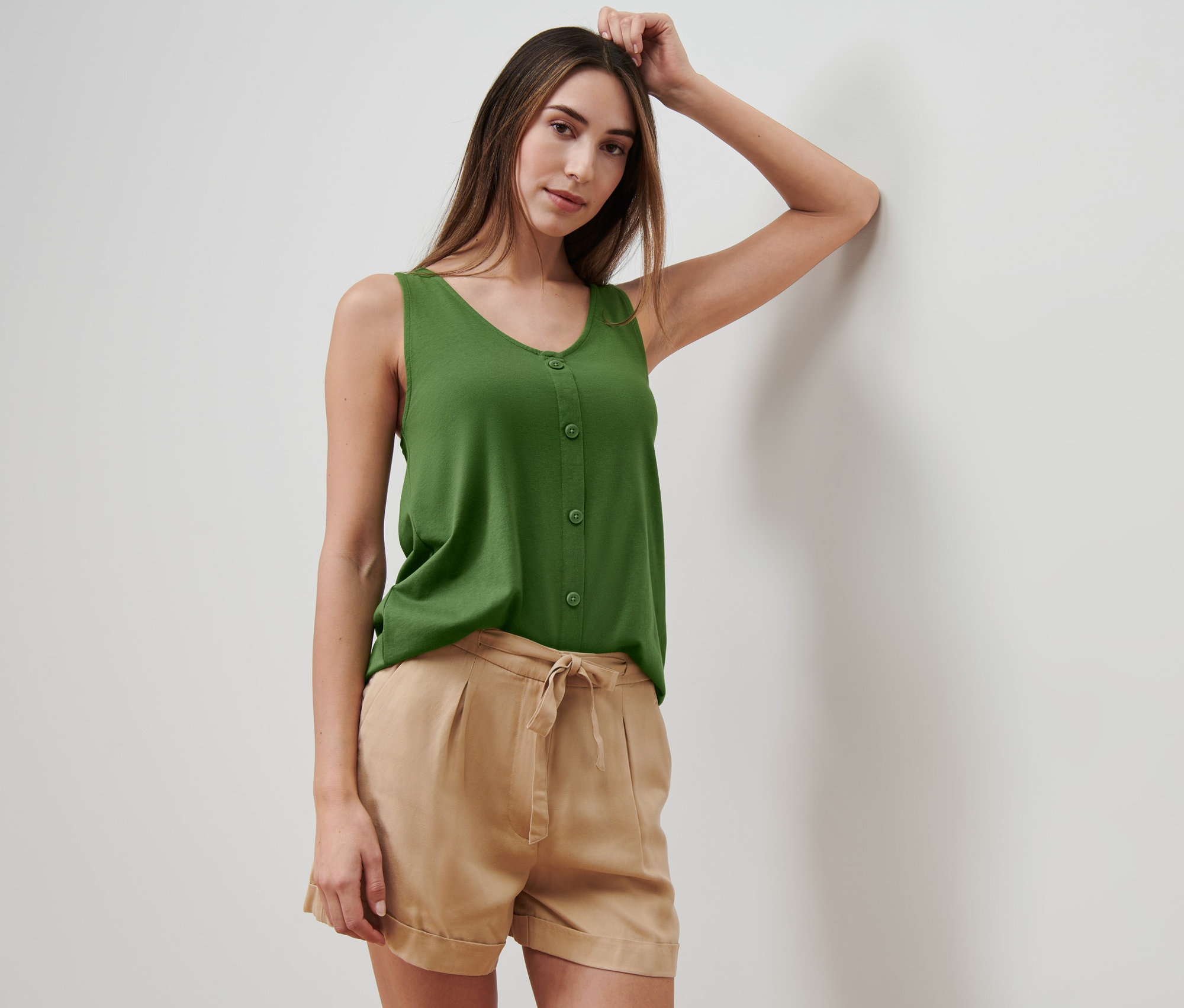 Femme appuyée contre un mur portant un haut vert avec patte de boutonnage et un short beige.
