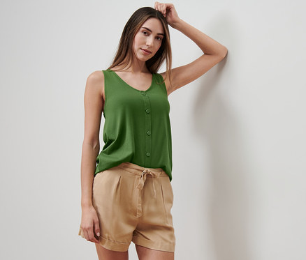 Femme appuyée contre un mur portant un haut vert avec patte de boutonnage et un short beige.