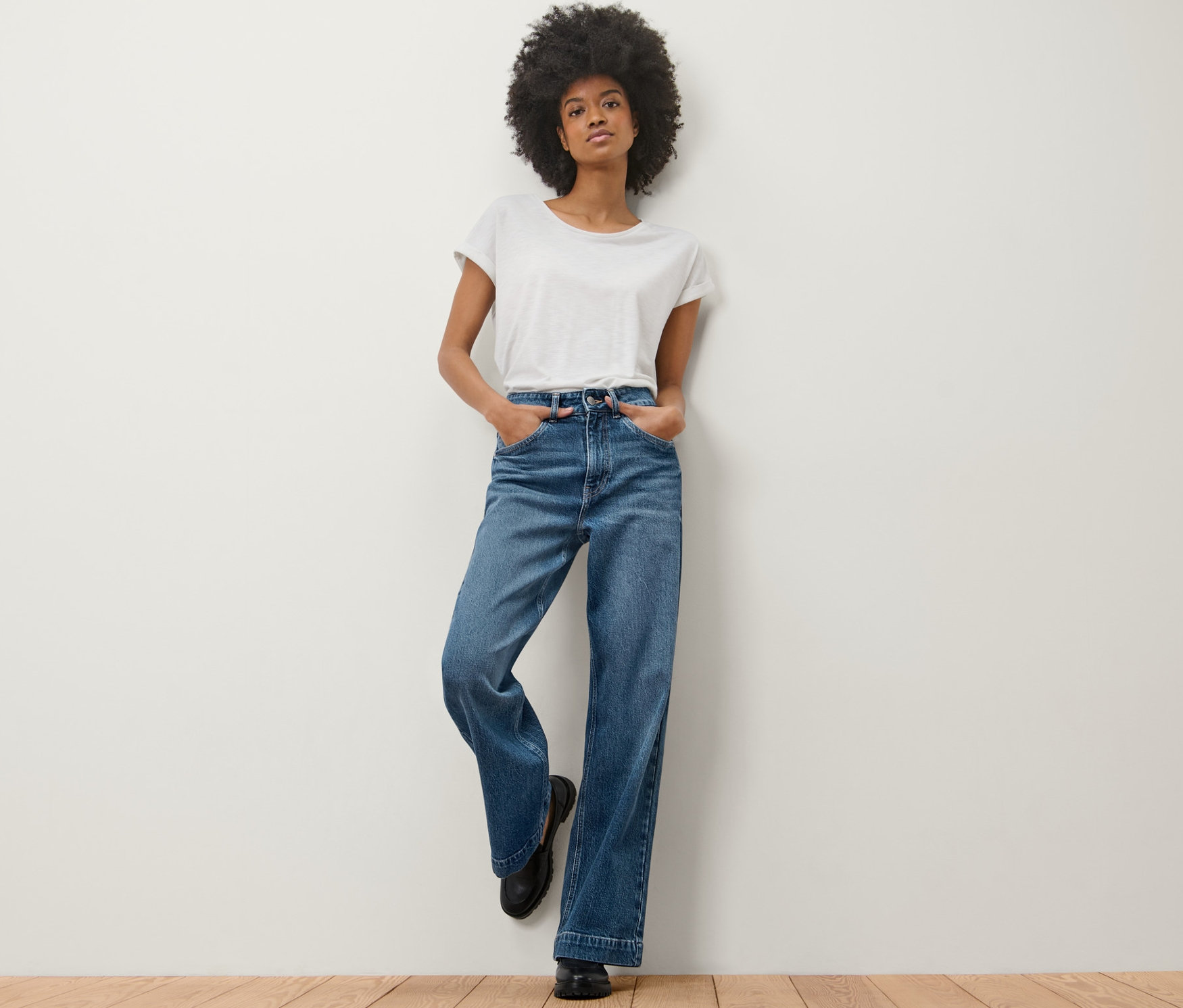 Une femme appuyée contre un mur porte un jean Wide Leg – Fit « Viki » et un t-shirt blanc.