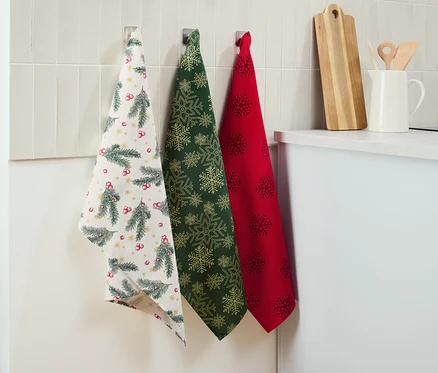 Trois linges de cuisine sont accrochés au mur : un blanc à motif de Noël, un vert à flocons de neige dorés et un rouge à flocons de neige foncés.