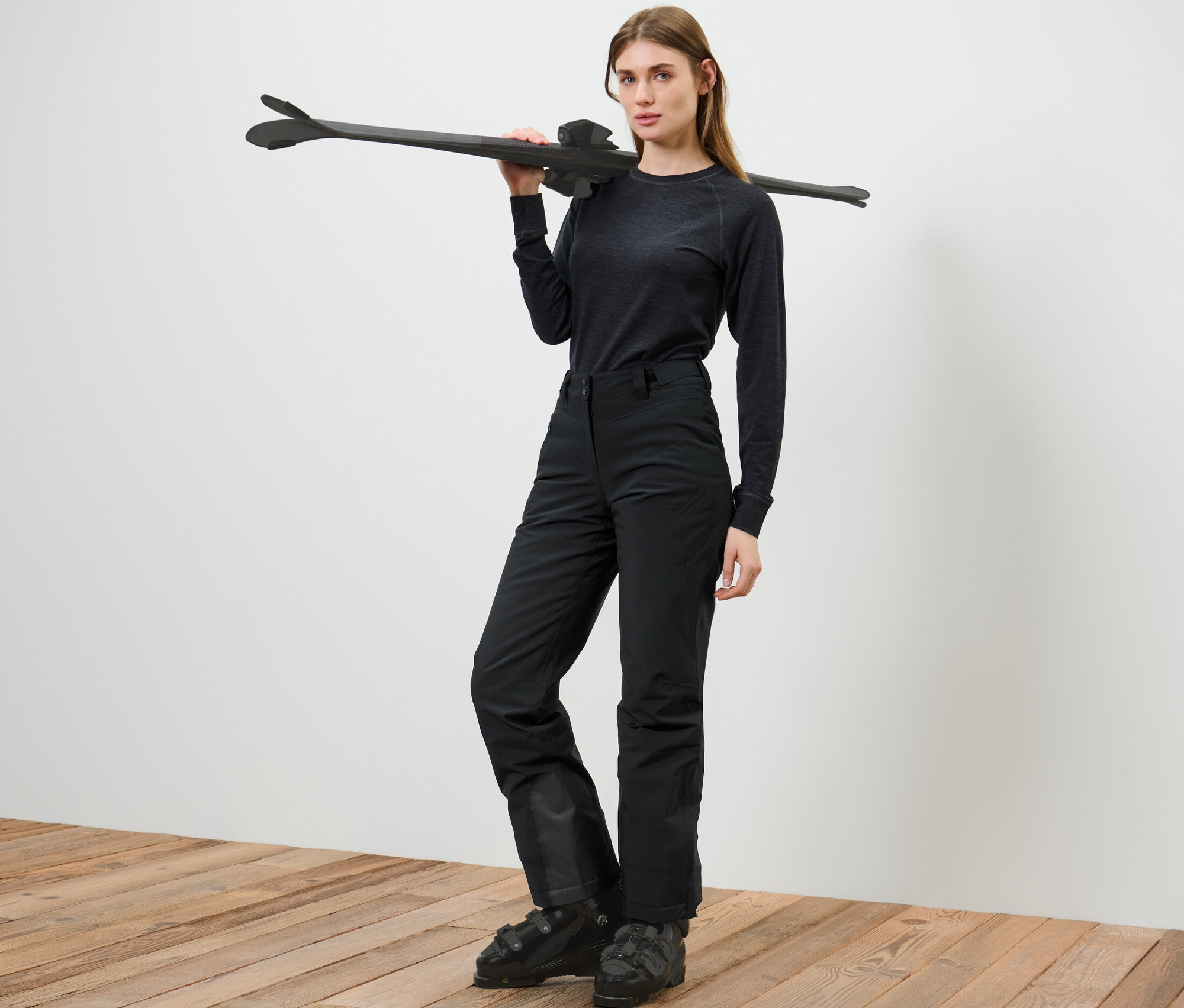 Femme posant en pantalon de ski, tenant des skis sur l'épaule.