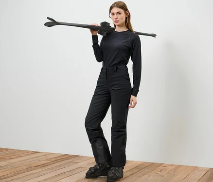 Femme posant en pantalon de ski, tenant des skis sur l'épaule.