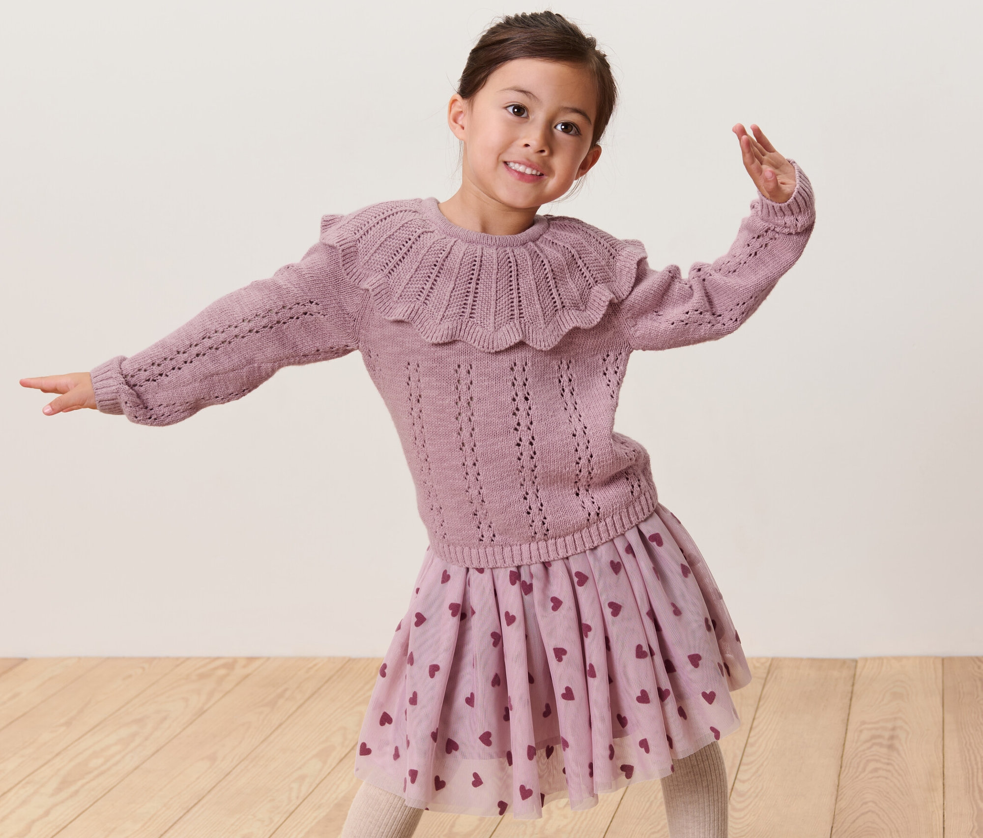 Une fille souriante pose, portant un pull en maille pour enfant avec col et une jupe à motifs de cœurs.