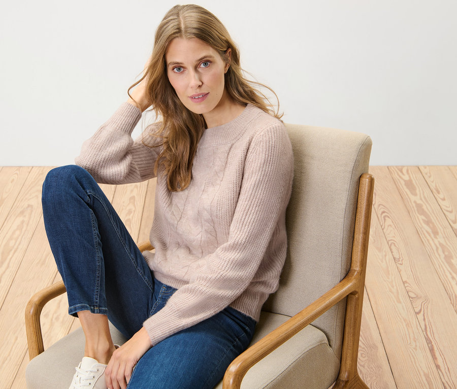 Femme assise dans un fauteuil portant un pull en grosse maille à motif torsadé.