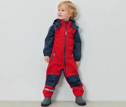 Petit garçon debout devant un mur blanc portant une combinaison en softshell pour enfant rouge et bleue.