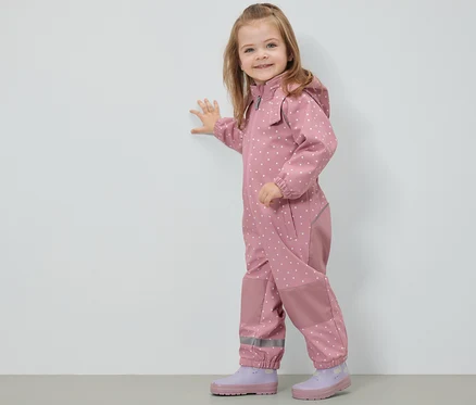 Une fillette souriante en combinaison en softshell pour enfant à pois se tient contre un mur.