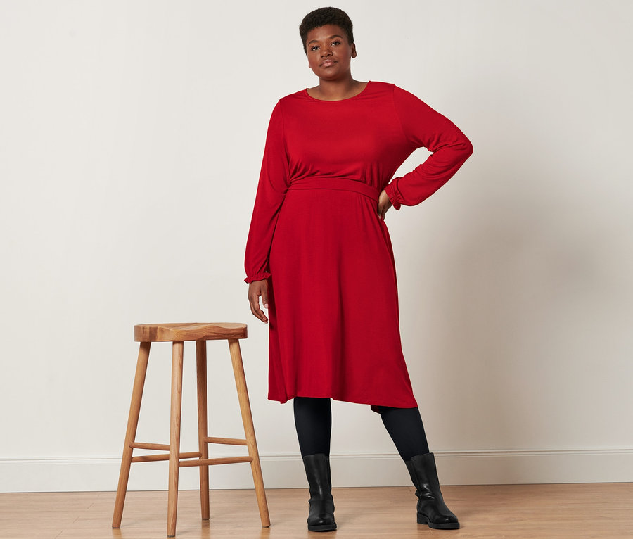 Une femme pose à côté d'un tabouret en bois et porte une robe en jersey mi-longueur, des bottines en cuir et 2 paires de collants opaques noirs.