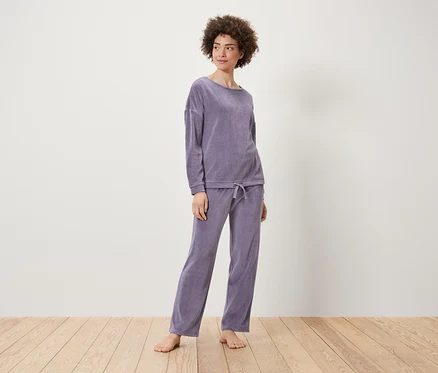 Une femme pose en ensemble en velours lilas composé d'un sweat-shirt et d'un pantalon sur un plancher en bois.