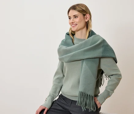 Une femme souriante porte un pull vert et une écharpe à carreaux avec des franges.