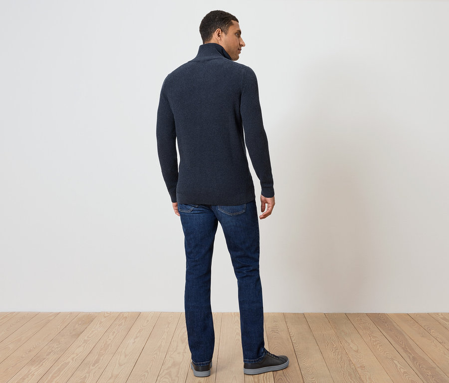 Un homme se tient dos à la caméra, portant un pull bleu foncé et un jean.