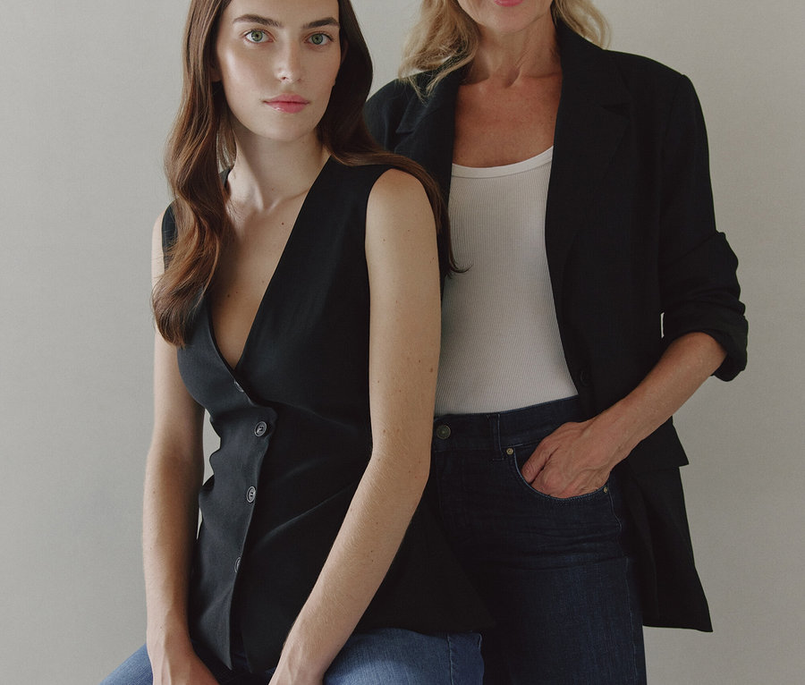 Deux femmes posent. L'une est assise sur un tabouret en haut noir et jeans, l'autre est debout derrière elle en blazer noir, débardeur blanc et jeans sombres.