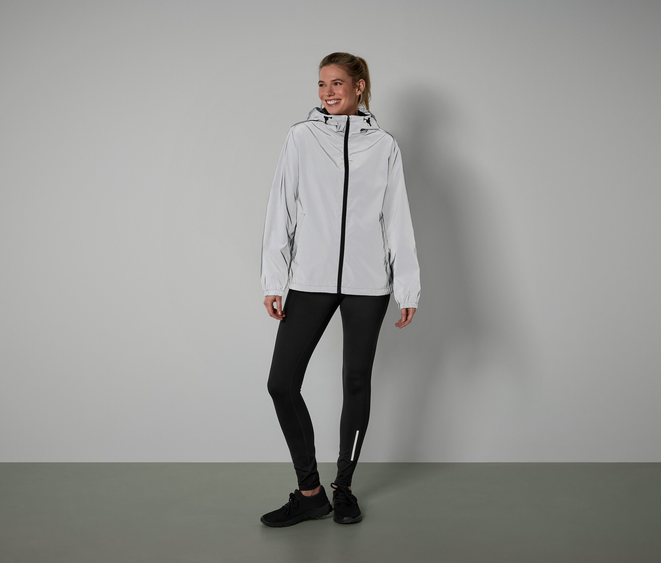 Une femme debout porte une veste argentée, un legging noir et des chaussures noires.