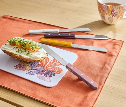 Sur un set de table orange se trouvent une tartine avec de la pâte à tartiner sur une planche à motif de feuilles, à côté se trouvent quatre couteaux avec des manches colorés et à l'arrière-plan une tasse de thé.