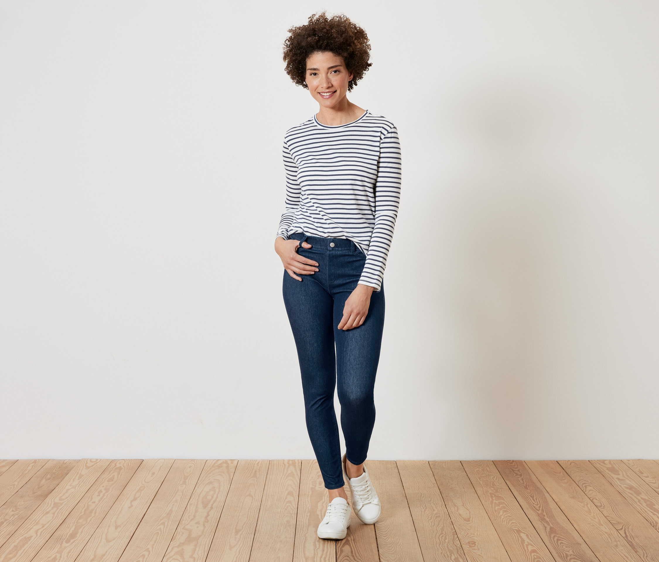 Femme aux cheveux bouclés posant dans un t-shirt rayé à manches longues, un jean bleu foncé et des baskets blanches.