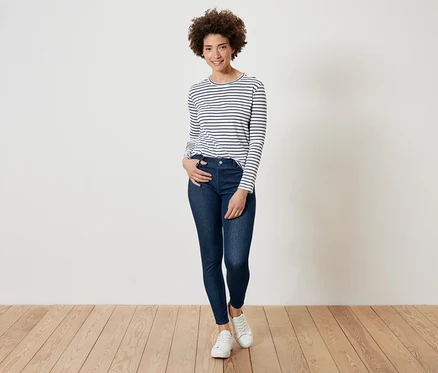 Femme aux cheveux bouclés posant dans un t-shirt rayé à manches longues, un jean bleu foncé et des baskets blanches.
