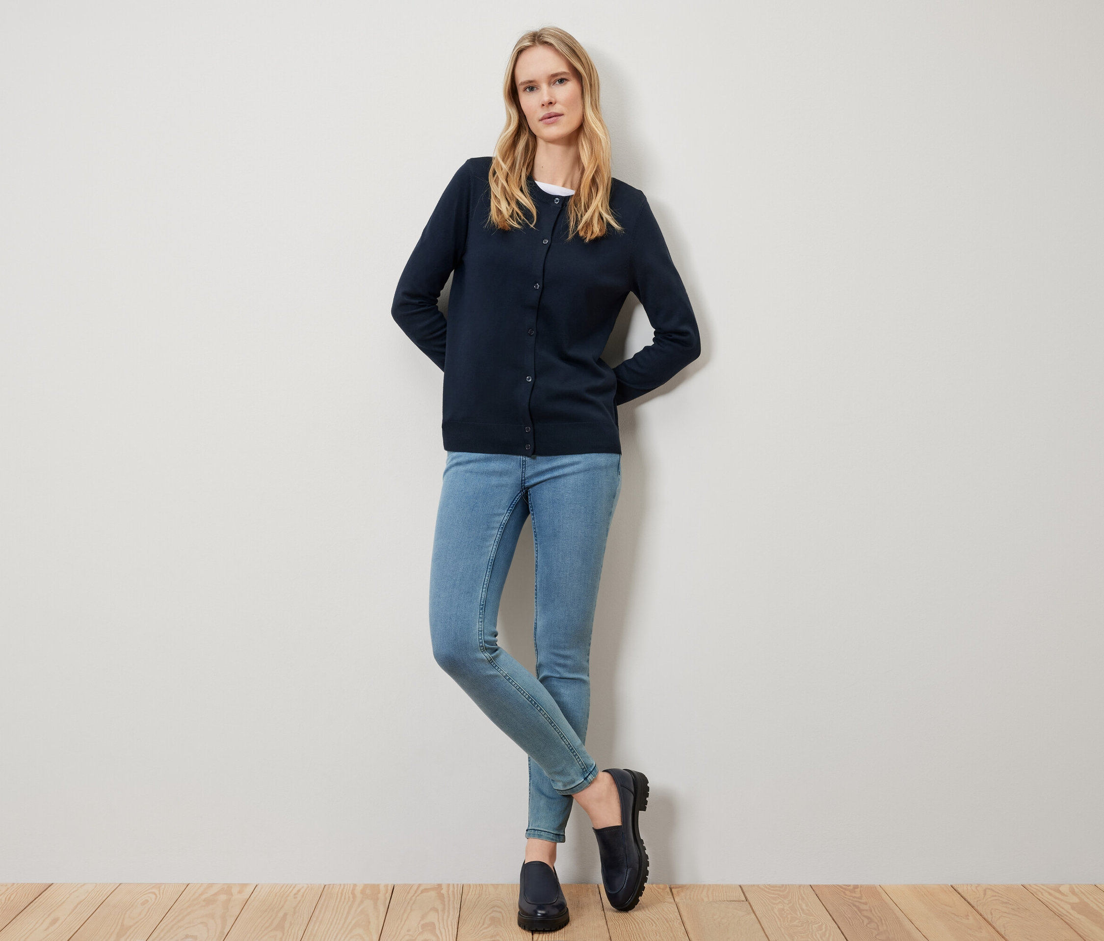 Une femme blonde portant un cardigan bleu marine, un jean et des chaussures noires pose devant un mur blanc.