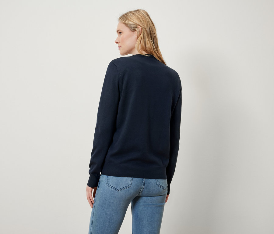 Une femme blonde, dos à la caméra, porte un pull bleu marine et un jean.