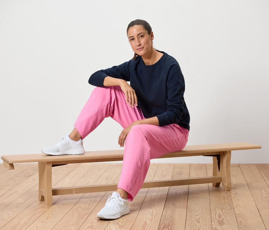 Une femme est assise sur un banc en bois, portant un sweat-shirt bleu marine, un pantalon rose et des baskets blanches.