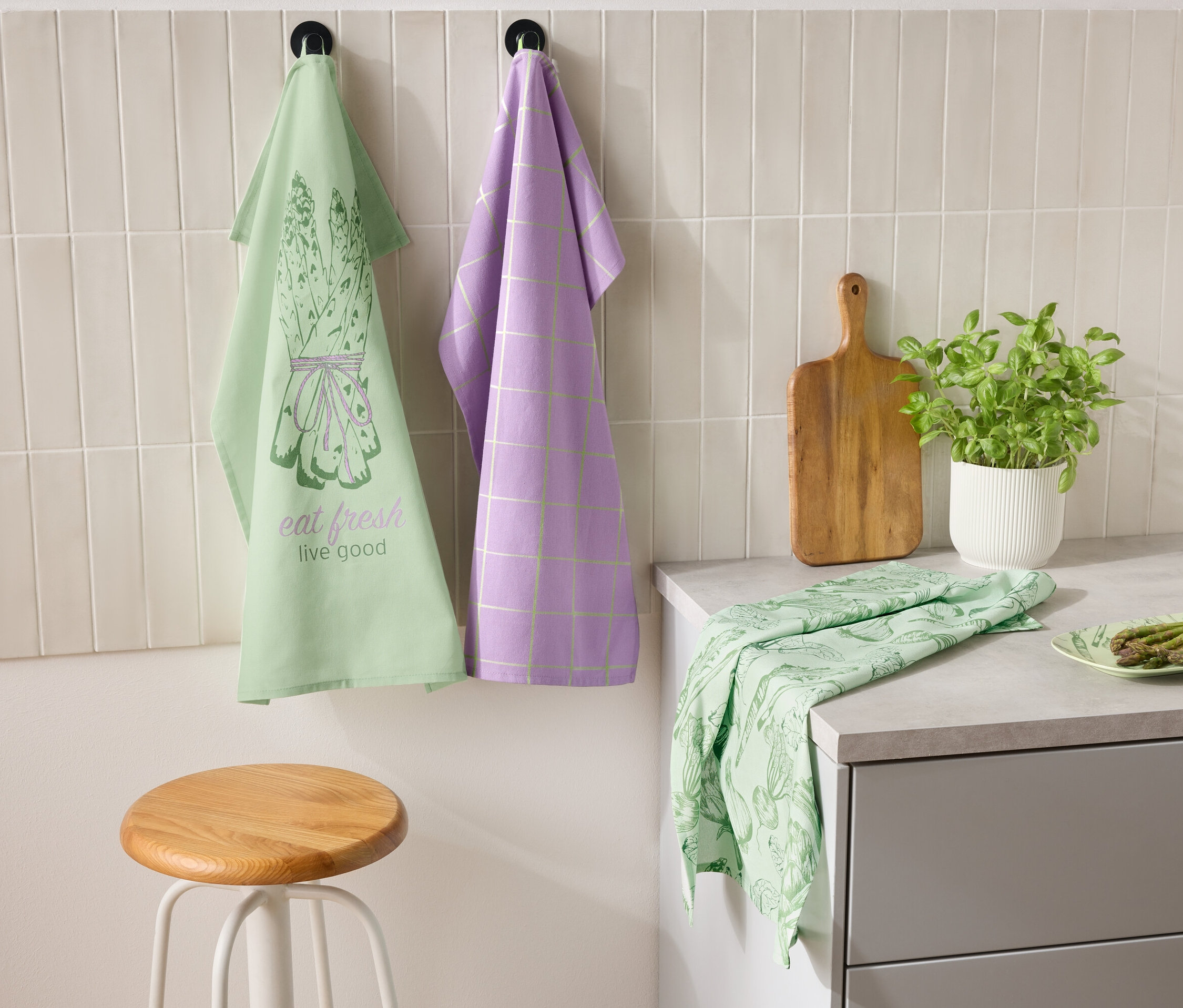 Deux torchons suspendus au mur côte à côte, un vert avec un motif d'asperges et l'inscription "eat fresh live good" et l'autre violet avec un motif à carreaux. Un torchon vert et une assiette d'asperges sont posés sur le plan de travail. À côté se trouvent une planche à découper et un pot de fines herbes.