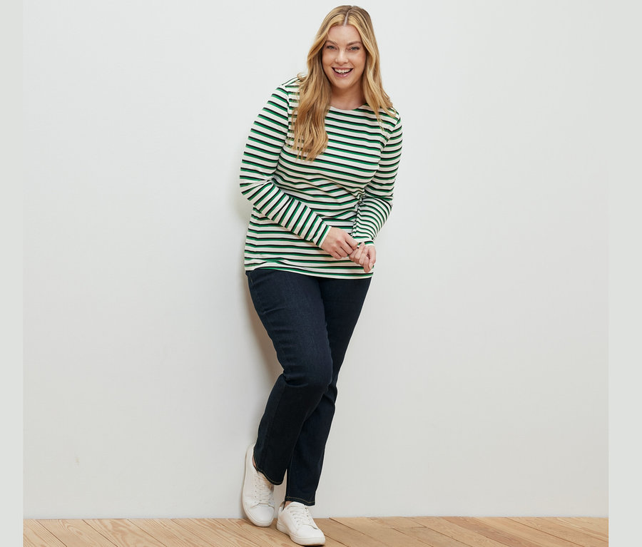 Une femme souriante appuyée contre un mur portant un haut à manches longues à rayures multicolores et un jean droit – Fit « Kira » bleu foncé.