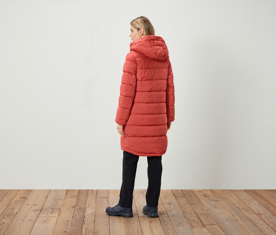 Une femme, vue de dos, porte un manteau matelassé rouge à capuche, un pantalon noir et des chaussures sombres.