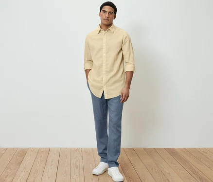 Un homme pose en chemise jaune en lin, pantalon bleu et chaussures blanches.