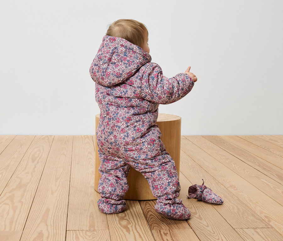 Un bébé est assis sur un tabouret en bois dans une combinaison à fleurs et pointe du doigt.