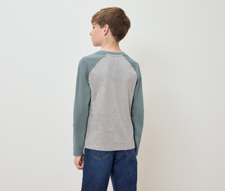 Un garçon se tient dos à la caméra, portant un t-shirt gris à manches longues vertes et un jean bleu.