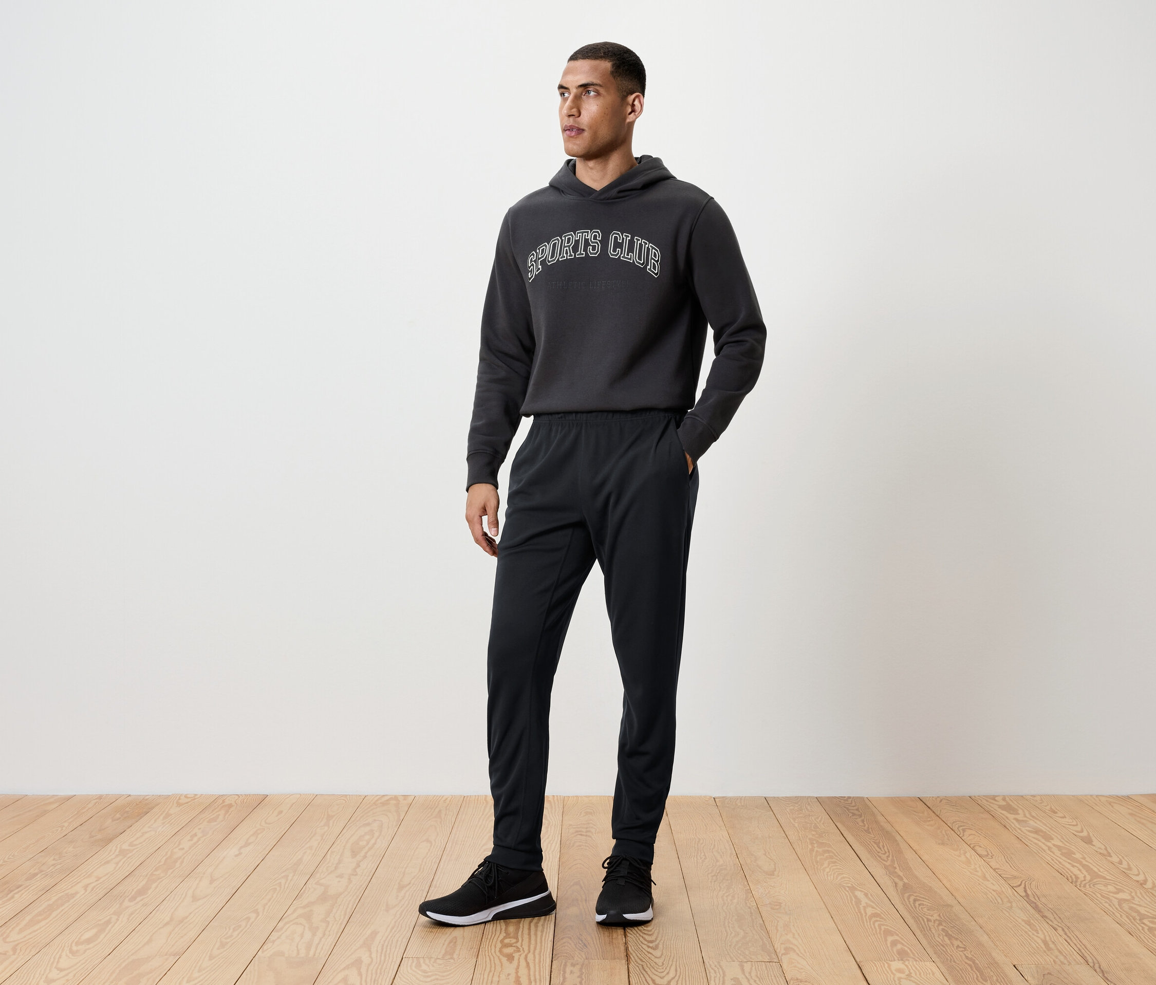 Un homme se tient debout sur un plancher en bois, portant un pantalon de survêtement noir, un sweat à capuche et des baskets.