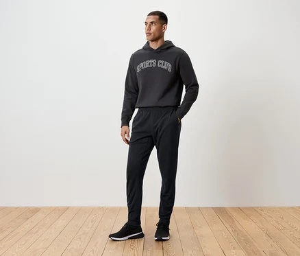 Un homme se tient debout sur un plancher en bois, portant un pantalon de survêtement noir, un sweat à capuche et des baskets.