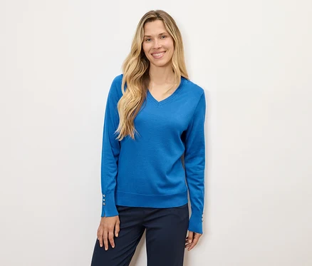 Une femme aux longs cheveux blonds pose en pull bleu et pantalon bleu marine.