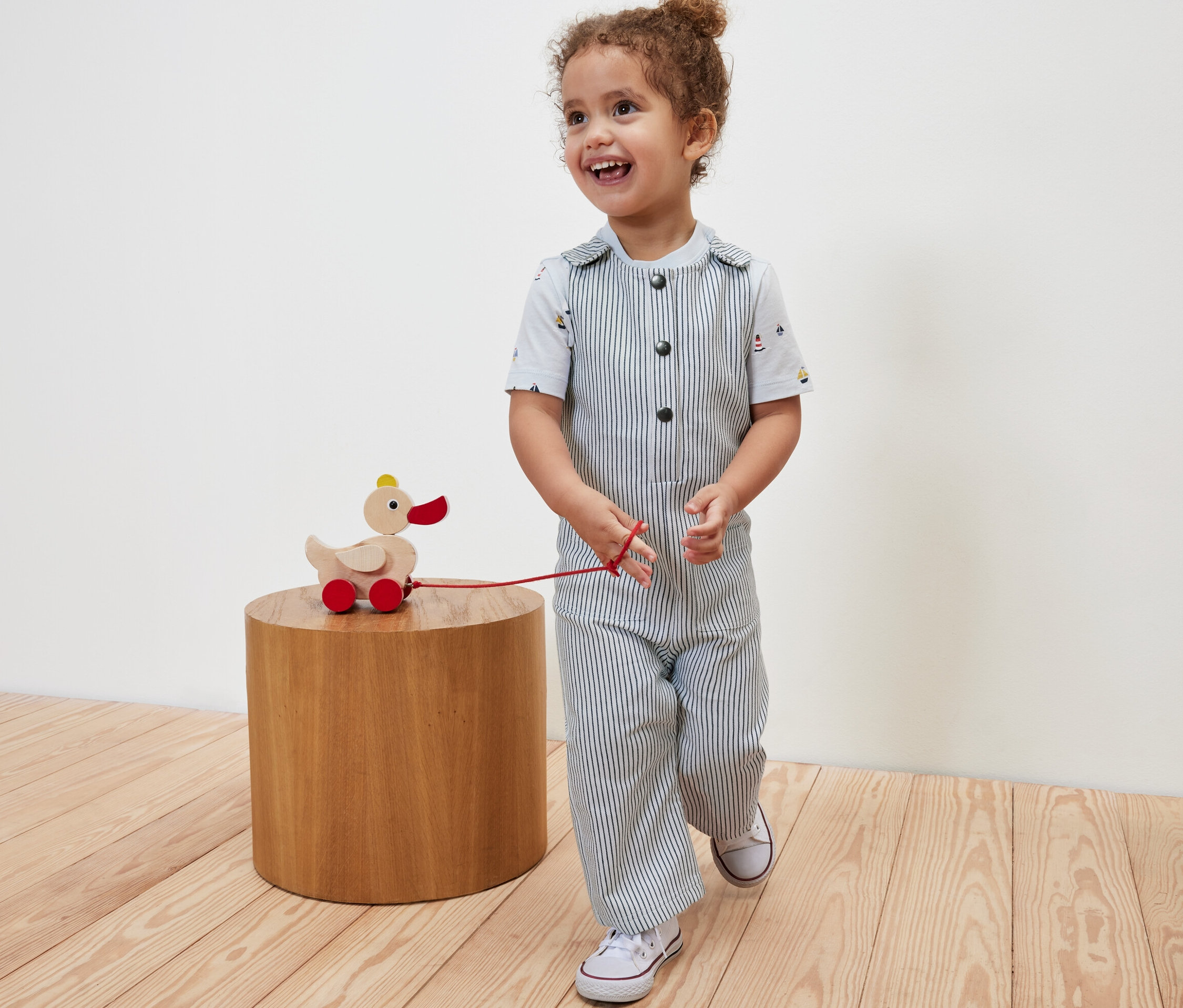 Enfant souriant tirant un jouet de canard en bois sur roues et portant une salopette tissée pour enfants.