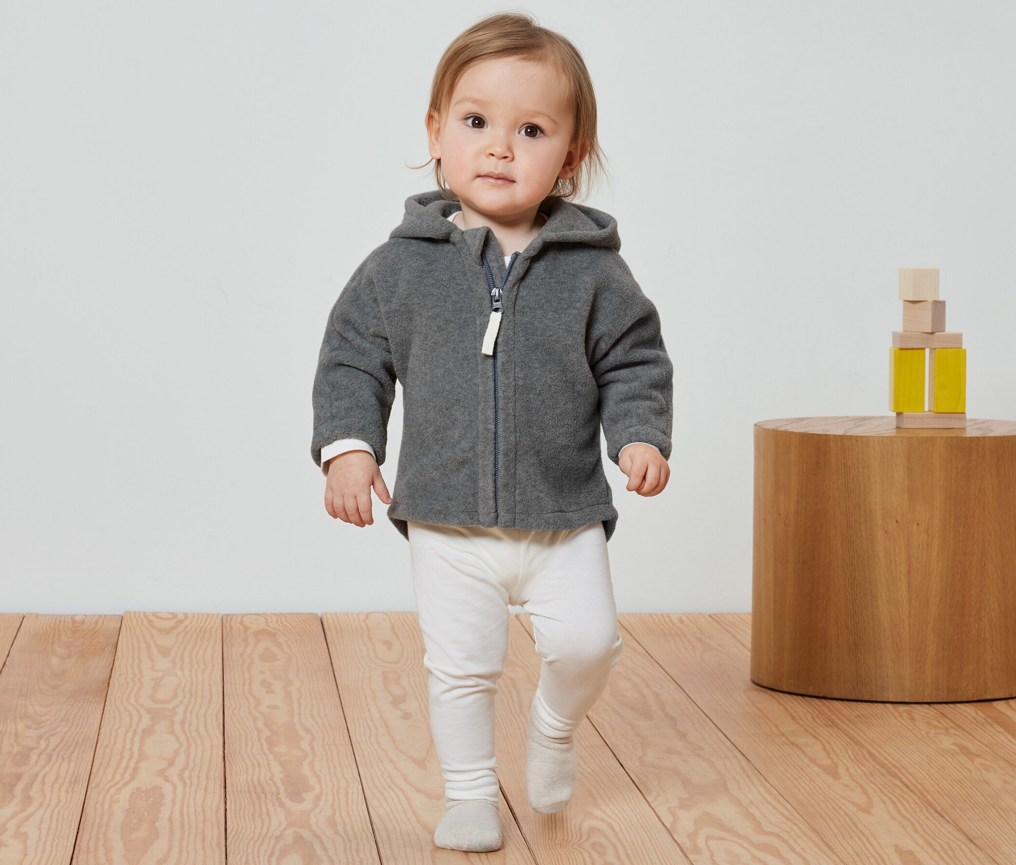 Un bébé marche avec une veste en polaire grise et un legging blanc.