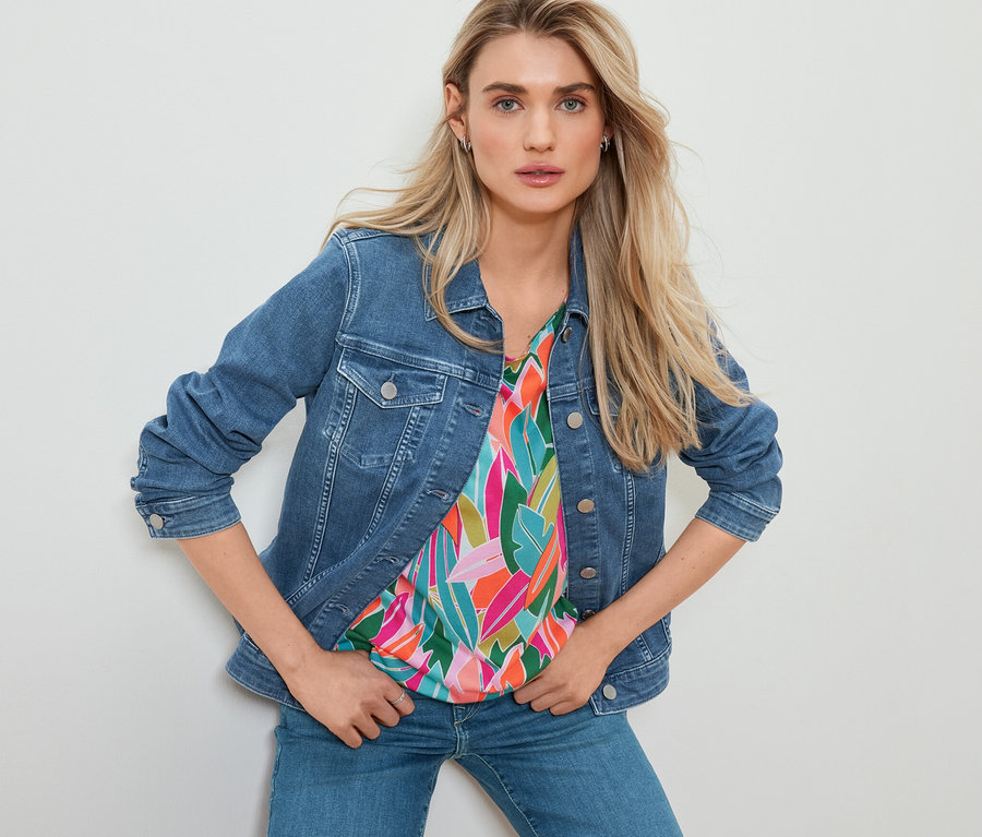 Une femme pose en veste en jean, un chemisier à encolure dégagée à imprimé intégral et un jean droit.
