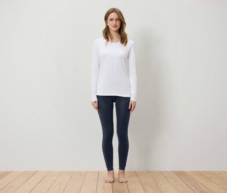 Une femme se tient sur un plancher en bois, portant un t-shirt blanc à manches longues et des leggings bleu foncé.