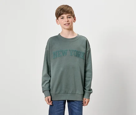 Un garçon porte un pull vert avec l'inscription NEW YORK et un jean bleu.