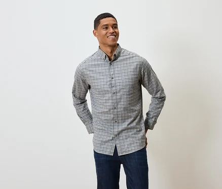 Un homme souriant pose en chemise grise à carreaux à manches longues et en jean bleu marine.