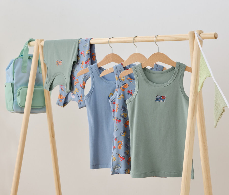 Des vêtements pour enfants sont suspendus à un portant en bois, notamment des débardeurs et un sac à dos.