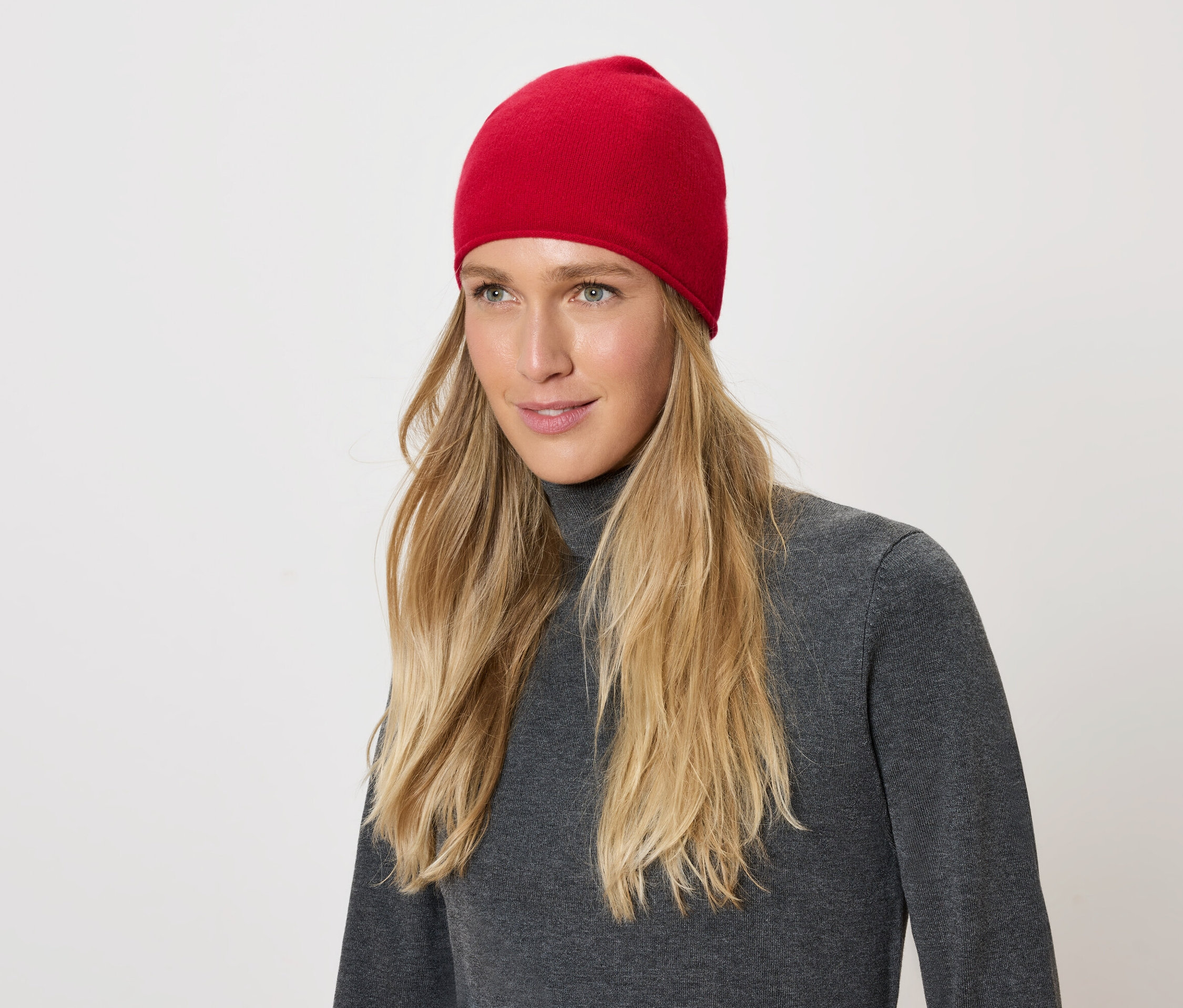 Portrait d'une femme aux cheveux blonds portant un bonnet rouge et un pull à col roulé gris sur fond blanc.