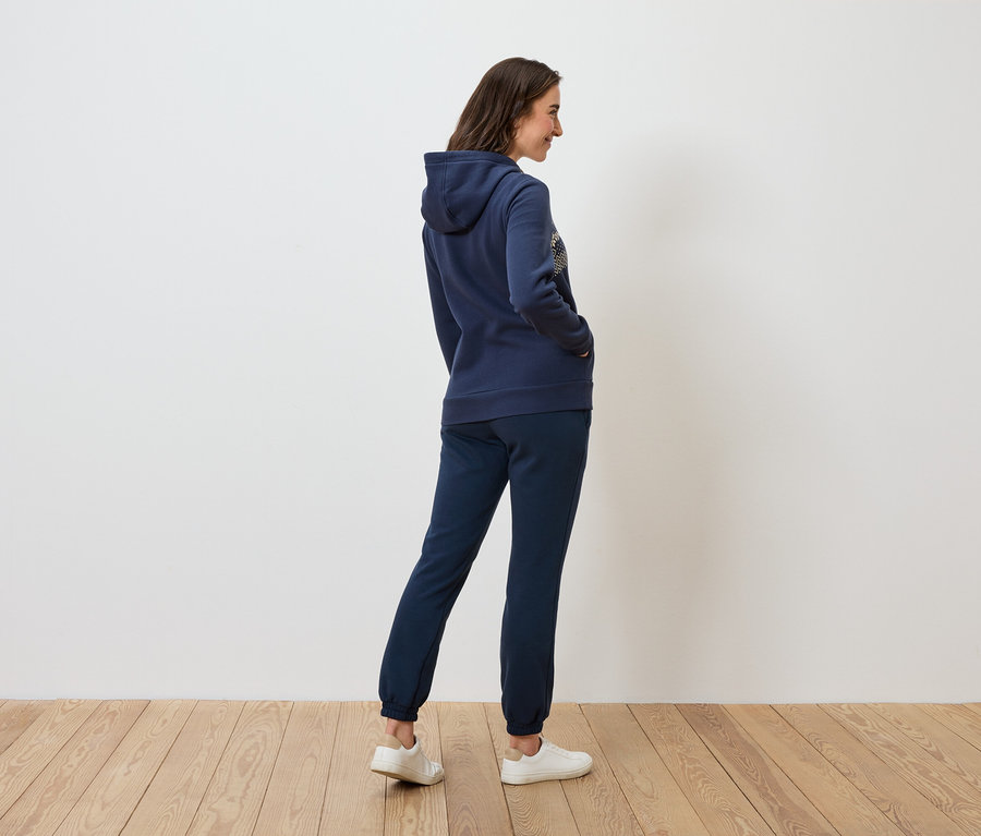Une femme portant un sweat à capuche bleu marine et un pantalon de survêtement se tient de côté sur un plancher de bois.