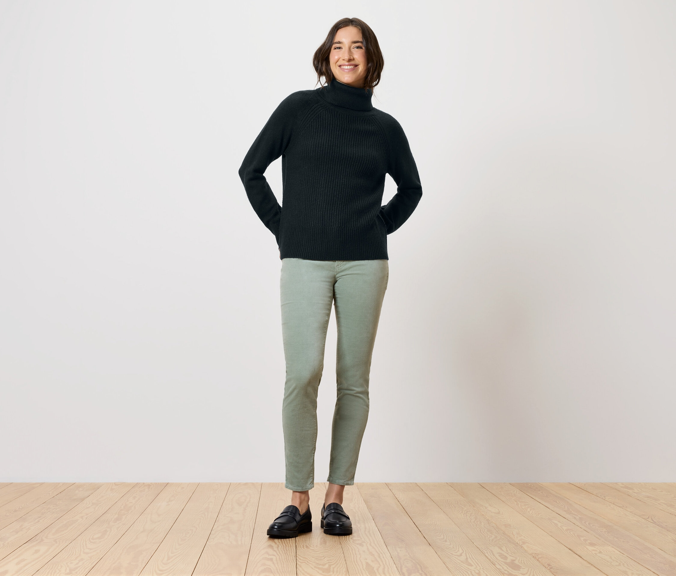 Une femme se tient debout, les mains derrière le dos, portant un pull à col roulé noir, un pantalon vert et des chaussures noires.