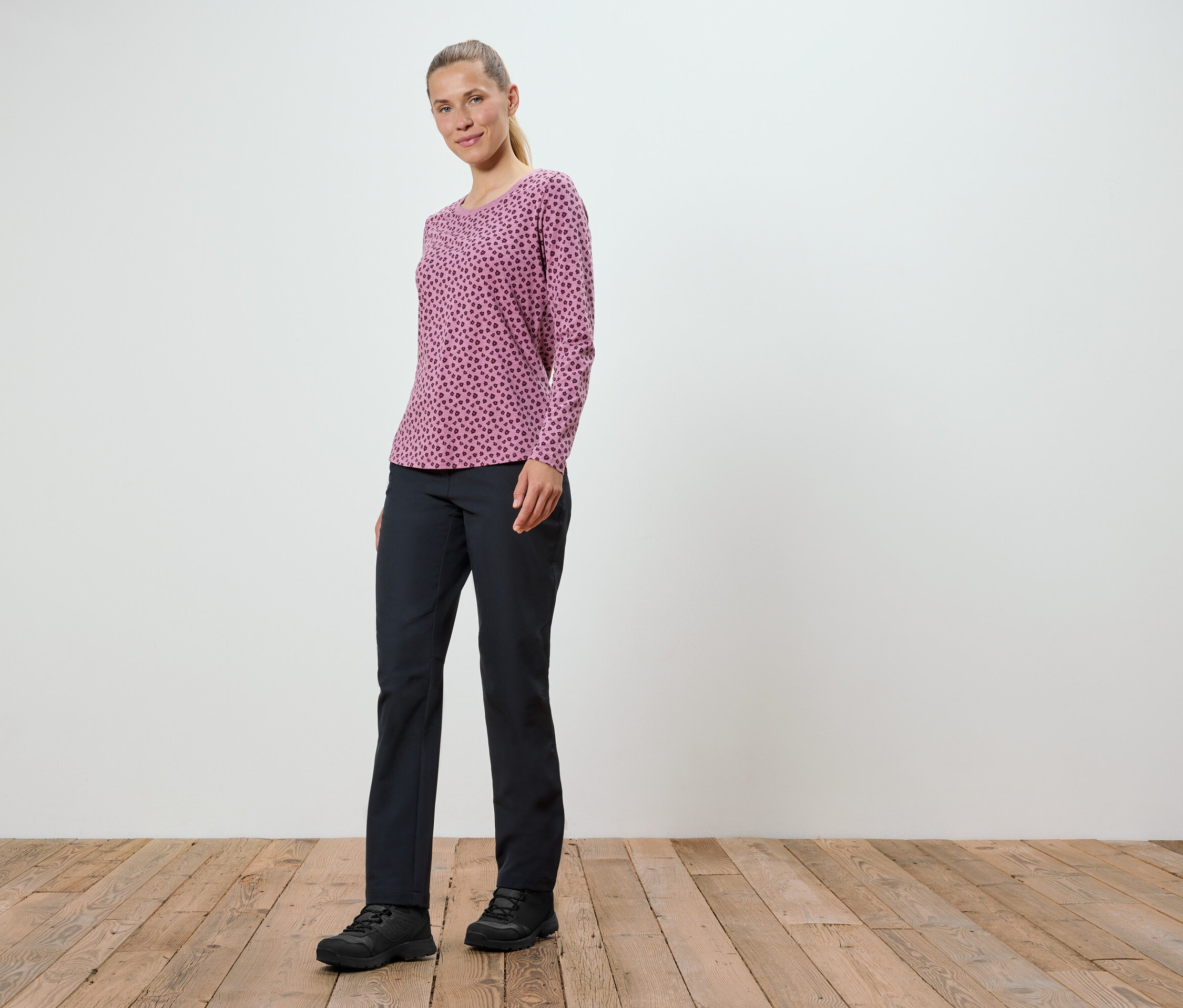 Une femme portant un t-shirt rose à manches longues et un pantalon noir se tient sur un plancher en bois.