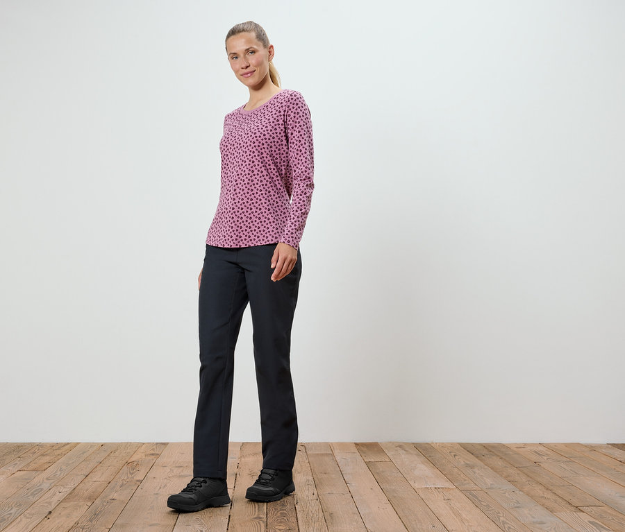 Une femme portant un t-shirt rose à manches longues et un pantalon noir se tient sur un plancher en bois.
